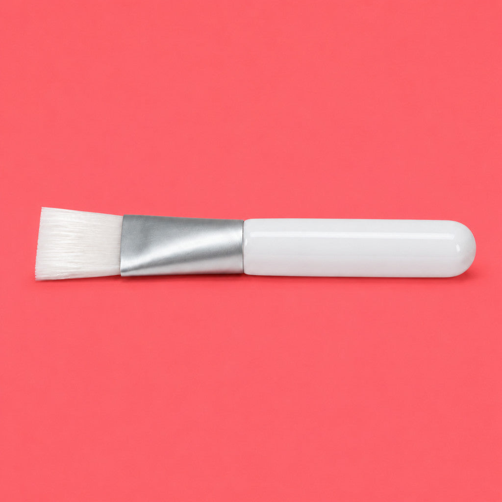 A skin care brush with white bristles, a silver metal ferrule, and a glossy white handle, positioned horizontally on a solid coral-pink background.
