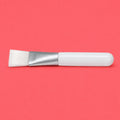 A skin care brush with white bristles, a silver metal ferrule, and a glossy white handle, positioned horizontally on a solid coral-pink background.